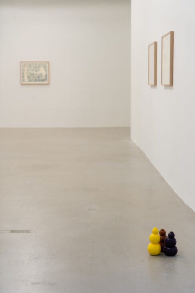 Vertical installation shot of "Kong Play" with three colorful Kongs clustered together in the lower right hand corner. Three drawings hang on the walls behind them, out of focus.