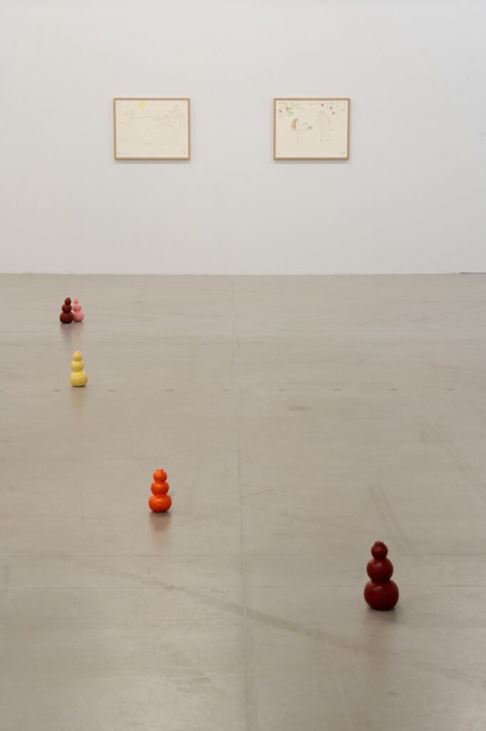 Vertical installation shot of "Kong Play" with a trail of brightly colored Kongs stretching from the foreground into the background. There are two drawings on the wall behind the Kongs.
