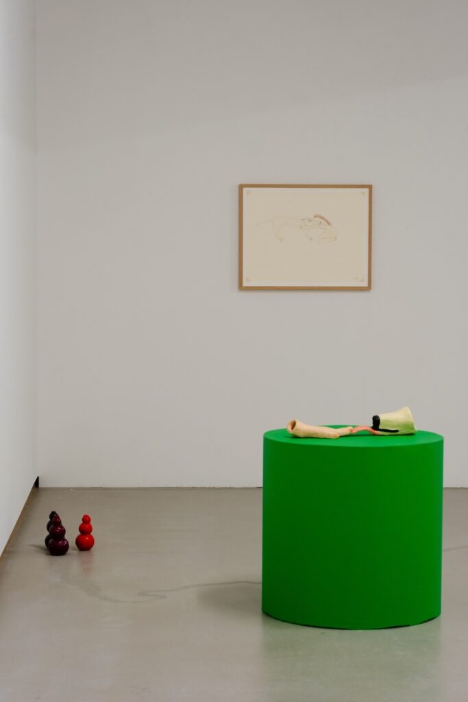 Vertical installation shot of "Kong Play" with three darker colored Kongs clustered in the lower lefthand corner. To their right, a ceramic human hand and dog snout with tongue extended rest upon a green plinth, the tips of the fingers rest on top of the tongue. A drawing depicting a long haired person hugging a Labrador Retriever hangs on the wall behind.