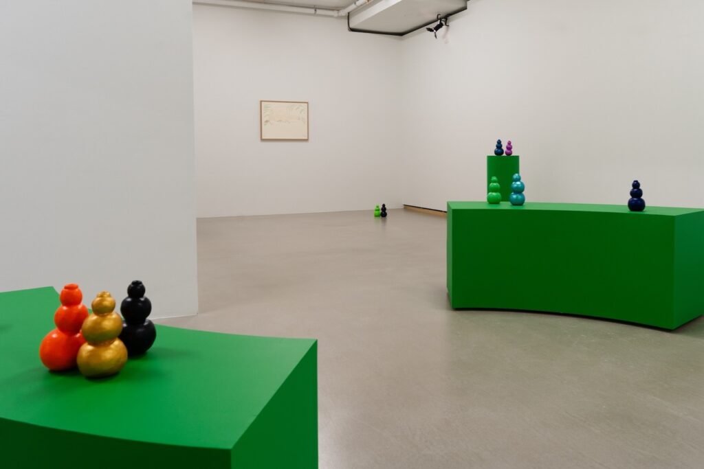 Installation shot of "Kong Play" with groups of brightly colored Kongs displayed on multiple green plinths throughout the room as well as the floor. There is a single, out of focus drawing on the back wall.
