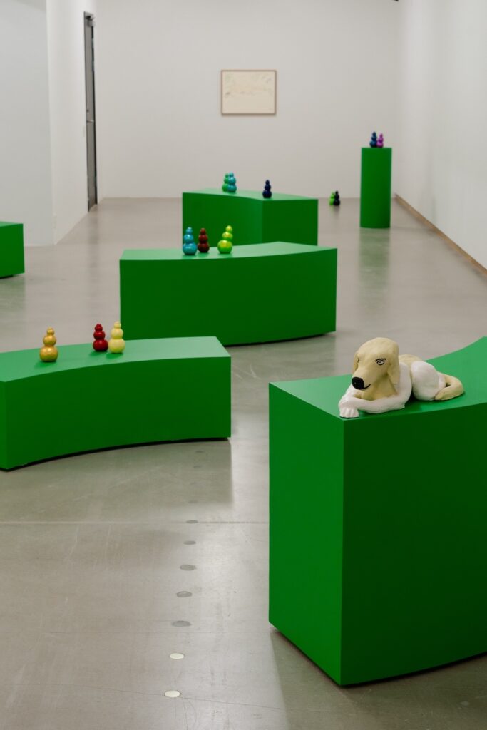 Vertical installation shot of "Kong Play" with a loose trail of green plinths extending backwards from the foreground. Kongs are displayed on all but the closest plinth, which instead has a ceramic sculpture depicting a blond sphinx-like Labrador Retriever dog with white, human arms and legs. There is an out of focus drawing on the back wall.