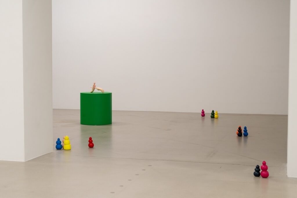 Installation shot of "Kong Play" looking through a large, doorless doorway. Pairs and trios of brightly colored Kongs are scattered across the floor. In the rear room, there is a single green plinth which holds a small sculpture depicting a pair of hands. One hand has an extended index finger which pierces through and comes out of the other side of the upturned palm of the second hand.