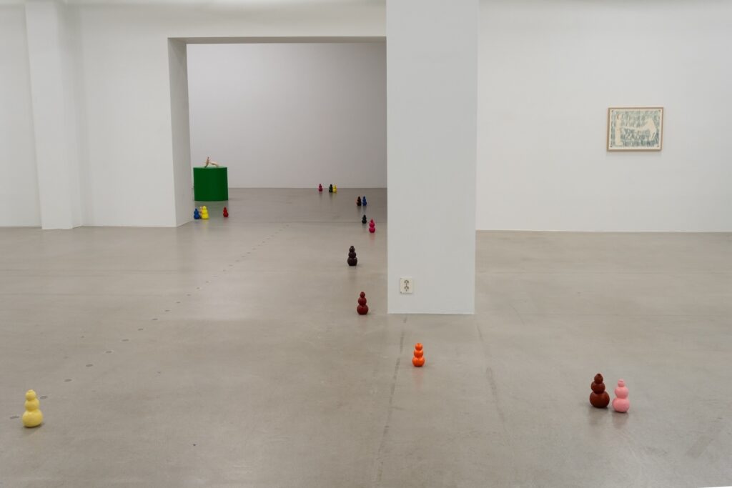 Installation shot of "Kong Play", taken a few steps further back from the previous image. A handful of more Kongs are visible on the floor in the foreground and to the right of the doorway, a single drawing hangs from the wall, depicting a woman and a Labrador Retriever facing each other against a dark blue background.