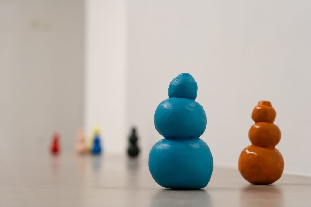 Installation shot of "Kong Play" depicting a turquoise Kong and a yellow-orange Kong standing side by side. A Kong vaguely resembles a three-tiered snowman or a sex toy. Some are ramrod straight while others are a bit lopsided. In the background, there are unfocused blotches of color indicating more Kongs.