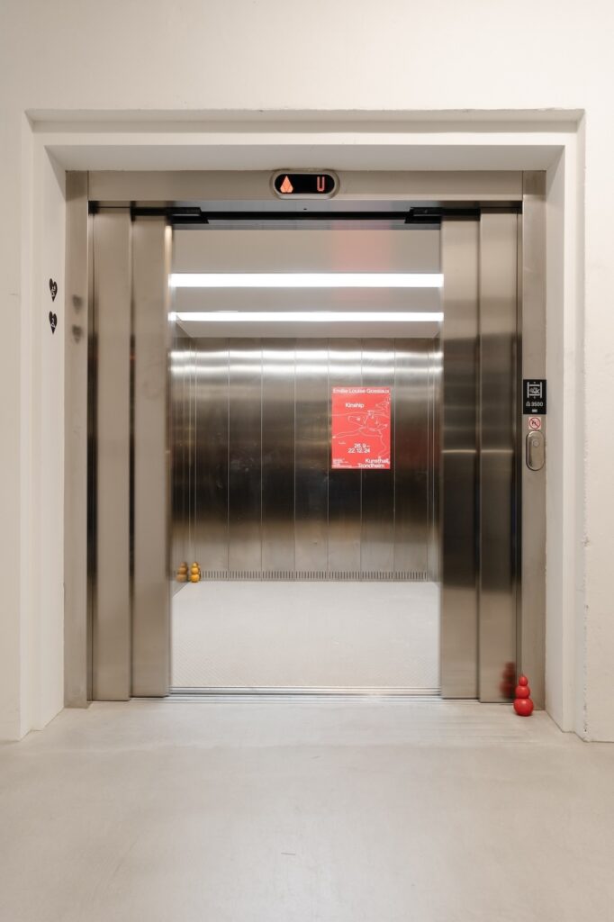 Vertical installation shot of "Kong Play" depicting the open silver doors of an elevator with a single red Kong placed right outside of the door and another gold Kong placed at the back left corner of the elevator.