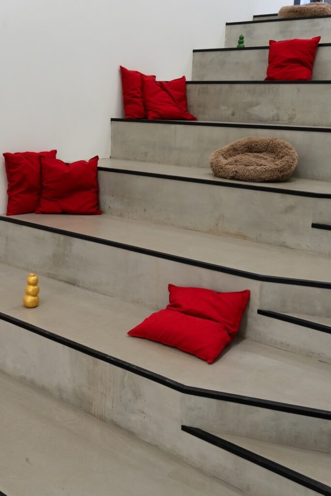 Vertical installation shot of "Kong Play" depicting large grey concrete steps with horizontal black edge guards. Bright red cushions and brown dog beds placed throughout. On the bottommost step, a single gold Kong stands alone. On the second to the top step, a forest green Kong is barely visible over the black lip of the step.