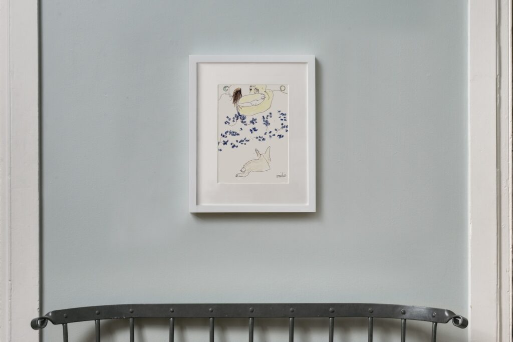 This is a vertical drawing in a white wooden frame hanging on a blue wall, called “”Love Bunny”. In this drawing, two people are lying in bed underneath a duvet patterned with vibrant blue flowers. At the foot of the bed, a small white bunny with a cotton tail is lying down facing away from the viewer. Above the heads of the two people in bed on the left side, is a small, blue crescent moon, and on the right side is a small yellow sun.