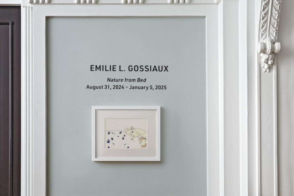 A landscape drawing in a white wooden frame hanging on a blue wall, called “Emilie and London in Bed”. A line drawing of Emilie (the artist) sleeping under a white duvet with blue flowers printed on it. London, Emilie's Yellow Lab guide dog, is curled up next to her head, also asleep.