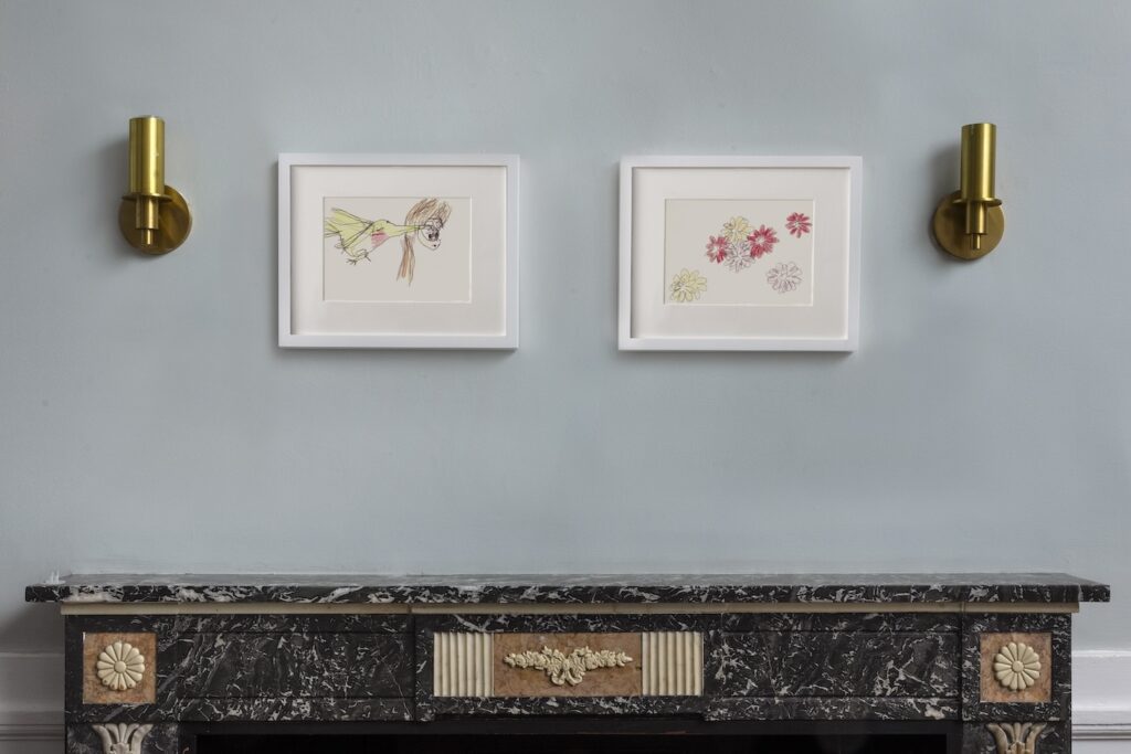 In this image, 2 landscape drawings in white wooden frames hang on a light yellow wall. On the left, is “Hummingbird in My Ear”. A blue green hummingbird with a red chin is poking her long beak out from the left side of the drawing. Its eyes are closed with long eyelashes, and her tongue curls out of her beak in a spiral, reaching into a human’s ear on the right side of the drawing. The ear has long brown hair tucked behind it. The second drawing on the right, is a landscape drawing, called “Flowers for London”, a drawing of a cluster of pink, scarlet, and peach flowers with 2 rows of petals circling around the center.