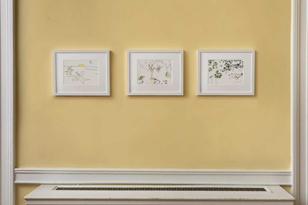 In this image, 3 landscape drawings in white wooden frames hang on a light yellow wall. The first drawing on the left is, “London Butterfly”. In this drawing, two English Labrador Retrievers with bright orange butterfly wings emerging from their backs are flying over a body of water. Over the water there is a setting Sun, and a crescent moon in the sky. The second drawing in the center, is called, “Greenhouse Flowers”. This is a line drawing of plants in Wave Hill's greenhouse. Small clusters of blue and purple flowers foreground the scene, with potted and hanging leafy tropical plants taking center stage. An arch of greenhouse windows and roofline frames the image at the top. And the last drawing on the right, is, London and I in the Meadow”. At the bottom right corner of the drawing, Emilie and her dog London are curled up together on the ground. Tall trees encircle them in the background, giving the impression of a forest.