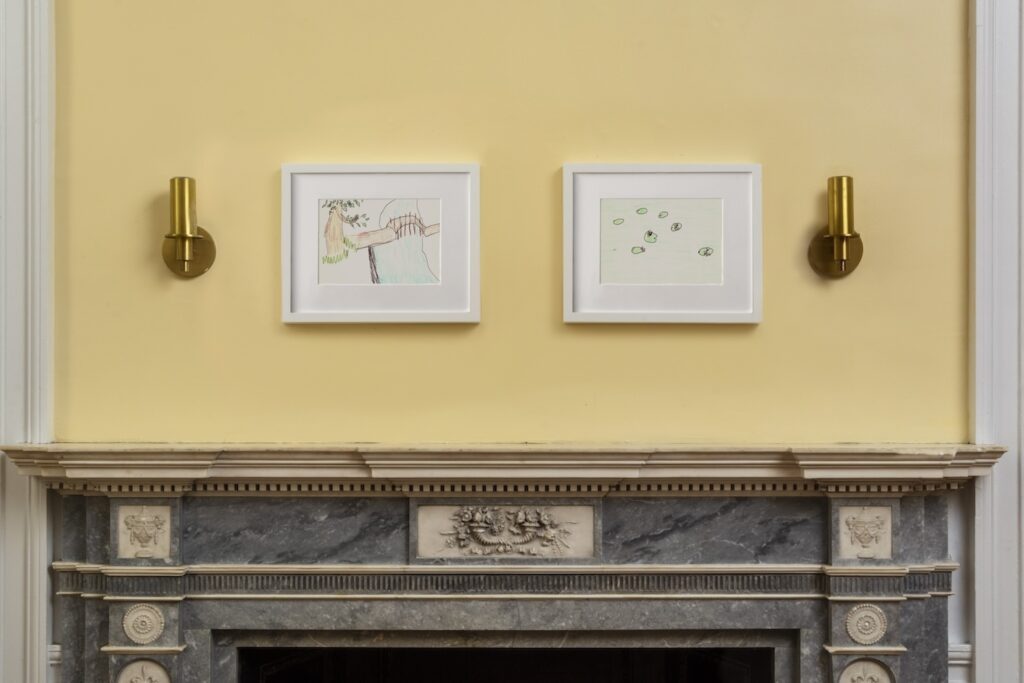 In this image, 2 drawings in white wooden frames hang on a light yellow wall. On the left, is a vertical drawing called “Bridge Over Stream”, in which a red wooden foot bridge stretches from the right side of the paper to the left side. Underneath it is a stream of turquoise blue water that stretches out towards the bottom of the paper. The drawing on the right, is a landscape drawing called, “Lily Pads in Water”, 7 green lily pads with light pink flowers float in a body of water that is turquoise blue.