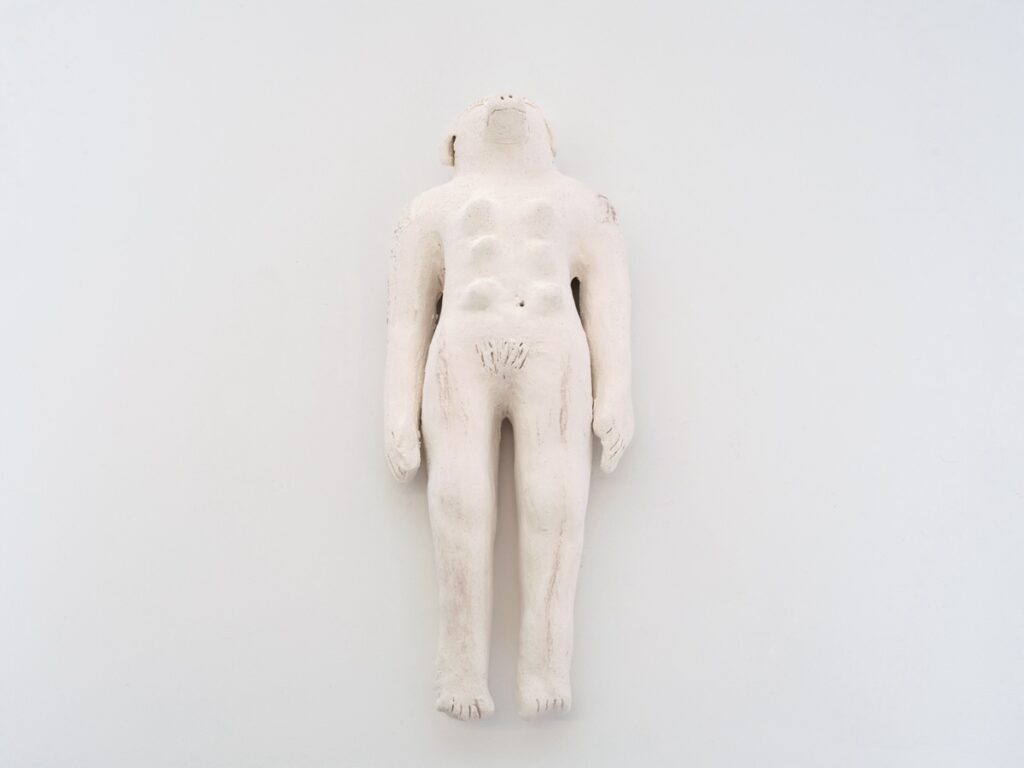 "Doggirl(larger)" is an off-white doll sized ceramic sculpture with a woman's body and a dog’s head and breasts. She is lying down on her back with her arms held closely against her sides. Her eyes are closed eyes and she has long eyelashes. She is lying on a white background.