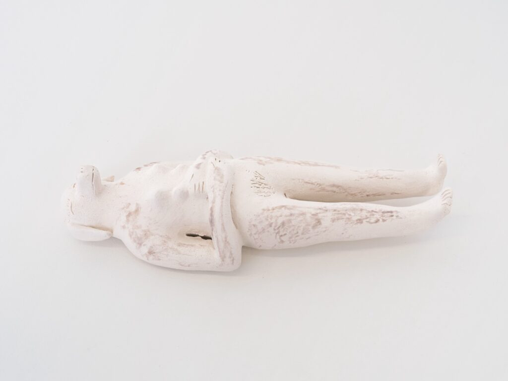 "Doggirl(smaller)" is an off-white doll sized ceramic sculpture with a woman's body and a dog’s head and breasts. She is lying on her back and her arms are folded 90 degrees at the elbows, with her hands resting on her stomach. Her eyes are closed eyes and she has long eyelashes. She is lying on a white background.