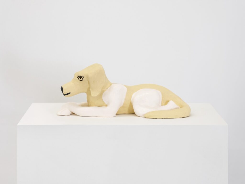 "Doggirl Sphinx" is a painted ceramic sculpture of a hybrid animal, half dog, half human, gently laying on the ground. Her form is inspired by the sphinx, a legendary ancient Egyptian creature representing strength and power. She has a dog's head and torso, but the arms and legs of a human. Her dog parts are painted blonde, while her arms and legs are painted white. Her face is painted with a kind and assured expression, featuring long black human-like eyelashes, while her human arms are folded over each other in repose.