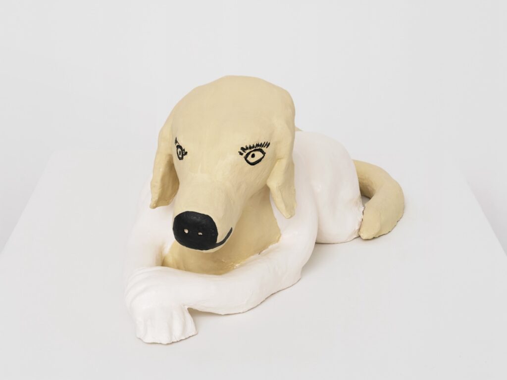 A detail view of "Doggirl Sphinx", focusing on her face. Her long, humanlike eyelashes are especially prominent. Beneath the carbon black tip of her snout, her bright white, human-style hands are folded neatly, one on top of the other.