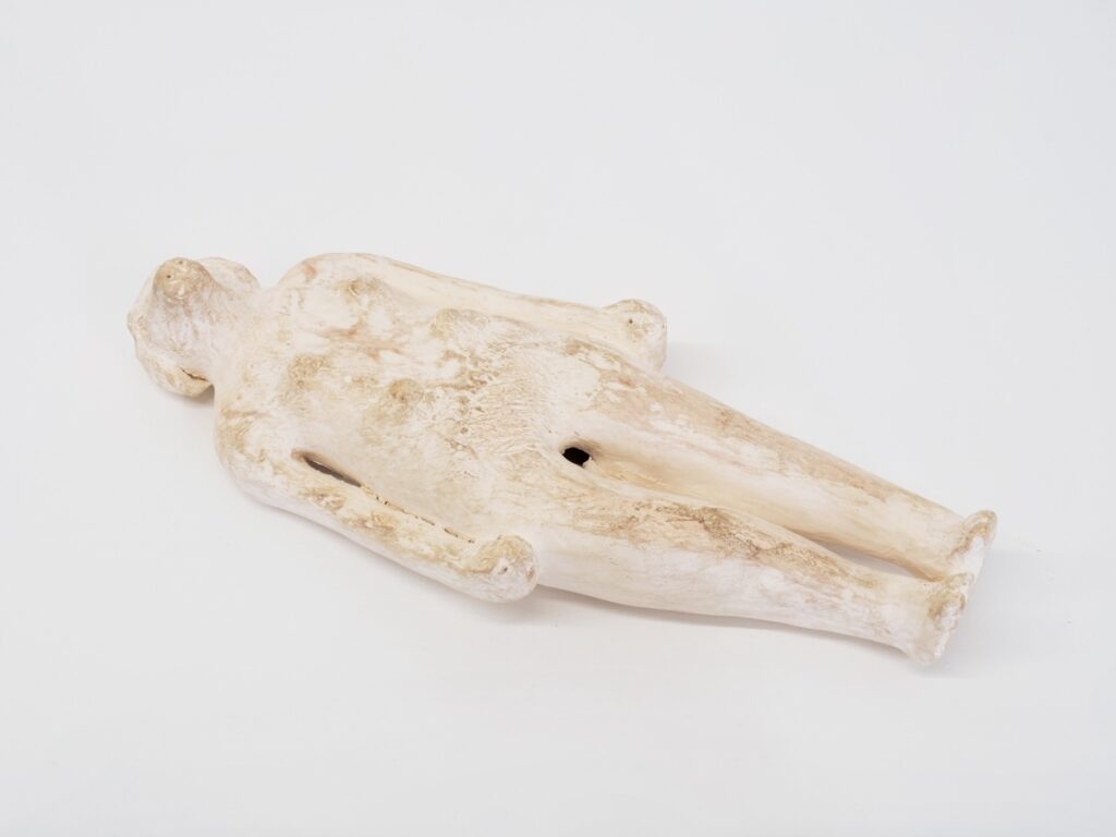 "Doggirl, They Called Me" is a white, doll-sized sculpture made from earthenware ceramic. The figure depicts a naked woman's body lying on her back with her arms held tightly against her sides. Instead of a human head, the artwork features a dog's head with long ears and her muzzle pointing to the sky. Additionally, her human torso has six nipples, resembling those of a dog.