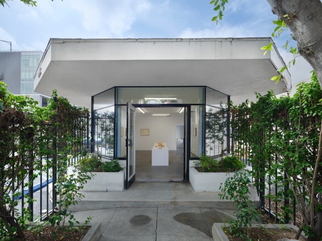 Exterior view of the show from the sidewalk in front of the gallery. A row of sculpture topped white plinths stretch deeper into the gallery, framed by the open doors. A flat, white roof hangs over the path that leads up to the doorway and two vine covered wrought iron fences parallel it. The scene feels serene.