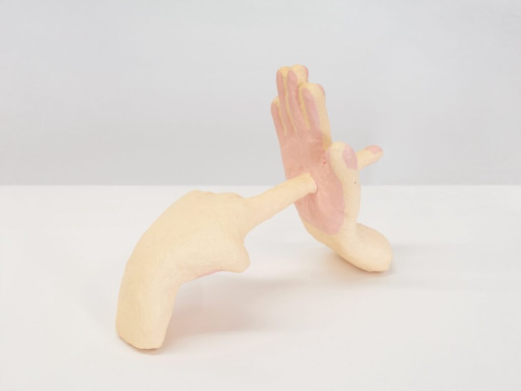 A detail view of “Finger Through Palm”, focused on the point where the extended finger pierces through the salmon colored palm.
