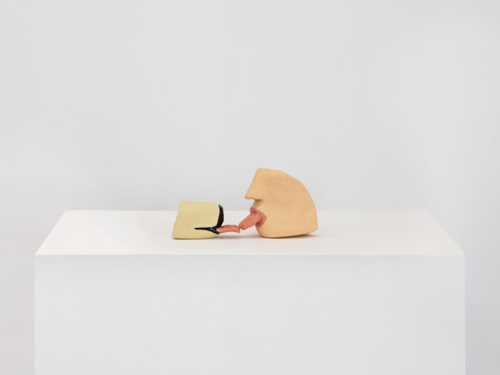 "French Kissing" is a painted ceramic sculpture made up of two parts. In this image, the part on the left is the blond snout of an English Labrador Retriever with its tongue stuck straight out. On the right, there is the lower section of a human face. The Human's tongue is also stuck out. The tips of the tongues touch.
