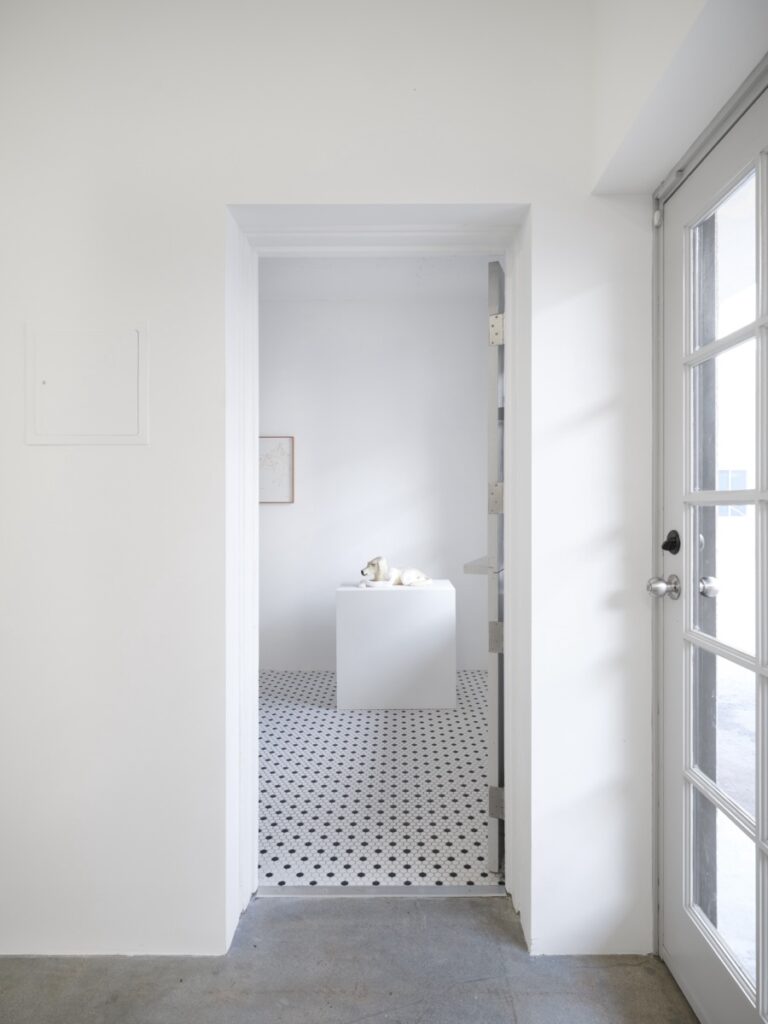 Vertical installation shot of "Doggirl Sphinx", viewed through a doorway. The sculpture and the white plinth that it sits on are framed perfectly by the open door.