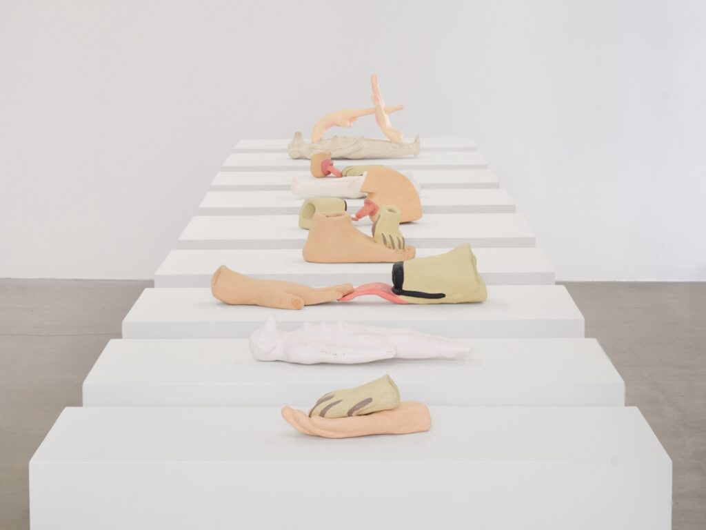 Installation shot of "Votives". A row of white, rectangular plinths stretches from the foreground backwards. Each plinth has a single sculpture on it. The plinths are perfectly aligned.