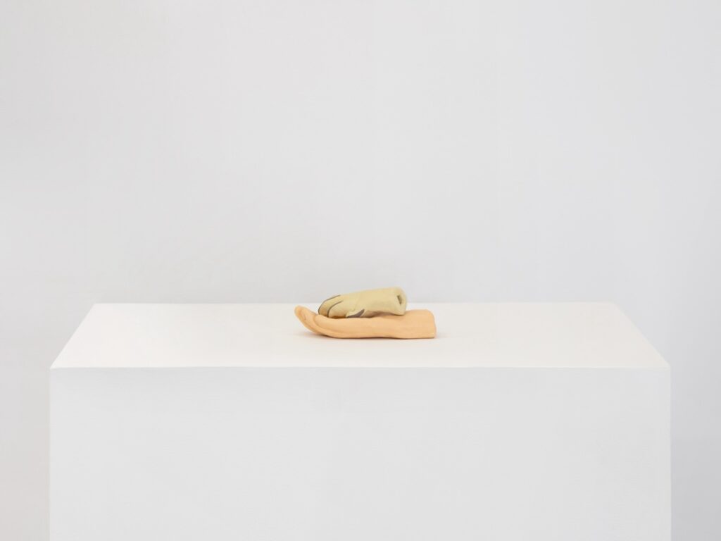 "Paw and Hand" is a painted ceramic sculpture made up of two pieces. On the bottom, an open human hand rests on the plinth palm side up. A blonde dog paw with grey-brown nails is nestled inside of the hand. Both the hand and the paw are cut off at the wrist.