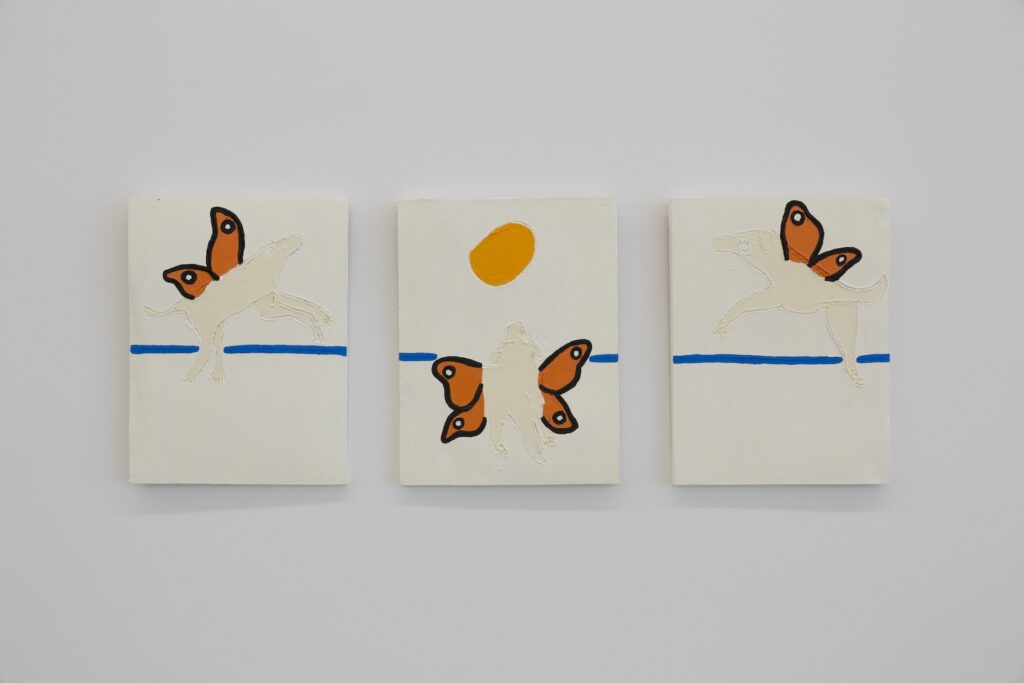 Three vertically-aligned rectangular tiles are arranged in a row on a wall. The tiles are cream colored and there is a single butterflydog on each. The butterflydogs look like English Labradors and have bright orange butterfly wings sprouting out of their backs with black and white highlights. The butterflydogs on the left and right tiles each point toward the center panel, giving us a profile view. The butterflydog in the center is viewed from behind, with her wings spread wide. She is looking up at a golden yellow sun above. A single blue line extends horizontally across all three tiles.