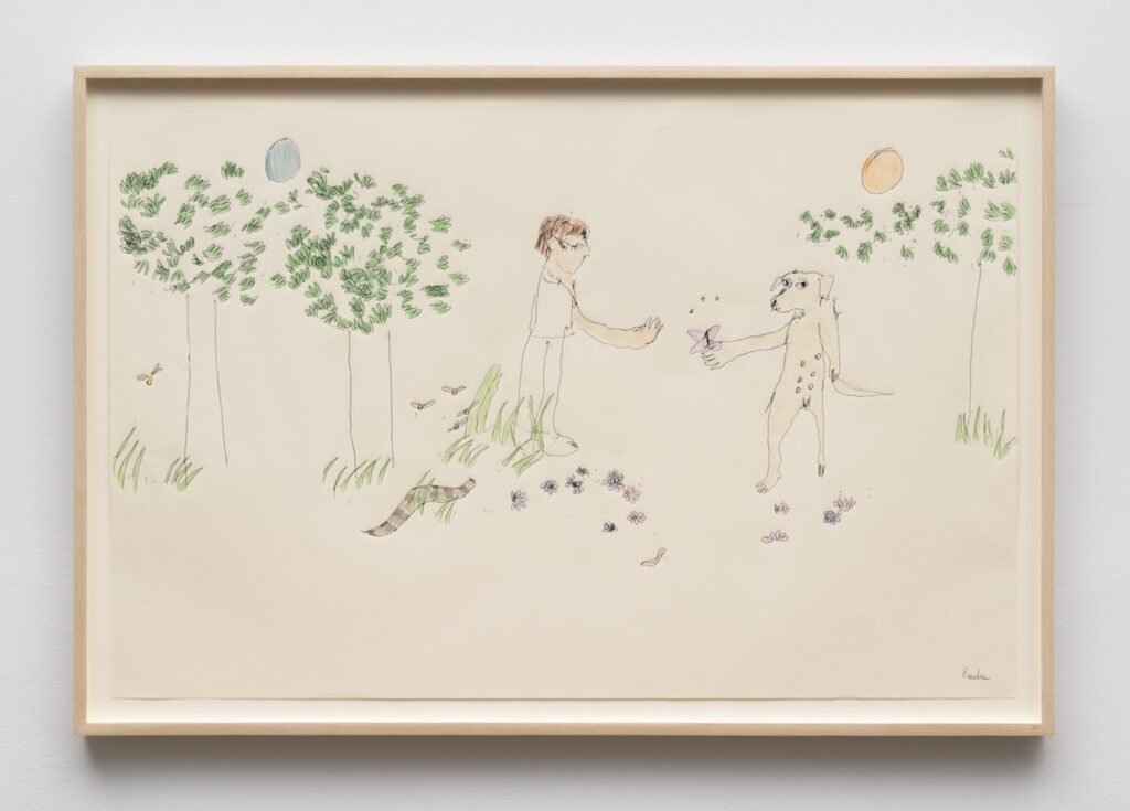 A childlike drawing on cream colored paper in a light wooden frame. The scene shows Emilie and London standing upright facing each other in the center, as if meeting in a clearing. Emilie has short brown hair, a simple pale outfit, and long, slightly exaggerated arms reaching toward London. London stands on two legs, has a long tail, and nipples on her torso, and is holding out a purple butterfly towards Emilie. Around their feet are small, loosely drawn flowers in purple and pink, and a tiny red worm-shaped line near the bottom. On the left side, three thin tree trunks support rounded clusters of green leaves, with a few tufts of grass and a brown, banded snake-like shape on the ground; small fire flies hover near the trees. On the right side, a single slender tree with a leafy green top stands near a patch of grass. There is a pale blue circle above the left trees and an orange circle above the right tree suggest a simple sun and moon in the sky. The background is otherwise blank, giving the drawing an open, airy feeling.
