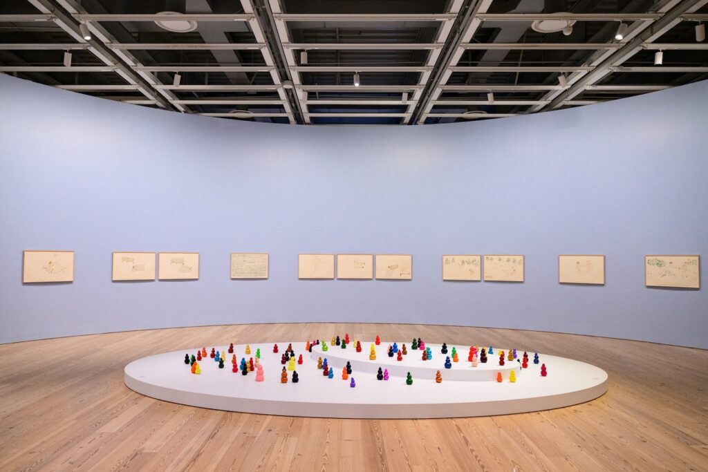 In the center of the gallery is my installation piece, “Kong Play”, which consists of 100 small ceramic sculptures of Kongs arranged in clusters and loose lines across a large, low, oval shaped platform. Each sculpture resembles three or four rounded shapes stacked vertically, like tiny abstract snowmen or butt plugs. They are painted bright, solid colors including red, orange, yellow, green, blue, purple, pink, and gold, creating a playful, colorful field against the white surface. The walls of the gallery are curved and painted pale blue, with 11 framed drawings on display.